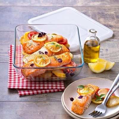 Travessa de vidro com salmão e rodelas de limão e tomate, prato com porção da comida, garrafa de óleo e fatias de limão sobre mesa de madeira.