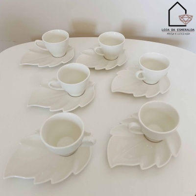 Seis chávenas de café brancas com pires em forma de folha sobre mesa branca com logótipo na parede.