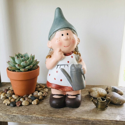 Figura decorativa de cerâmica de rapariga com chapéu verde e regador, vaso com suculenta e pedras sobre madeira