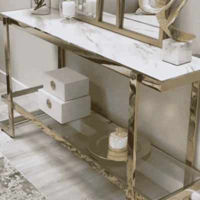 Consola com tampo em mármore branco e estrutura dourada, com caixas brancas e decoração sobre prato espelhado.