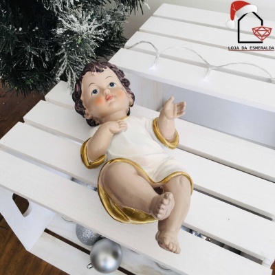 Boneco decorativo do Menino Jesus em resina com túnica branca e dourada sobre superfície branca de madeira