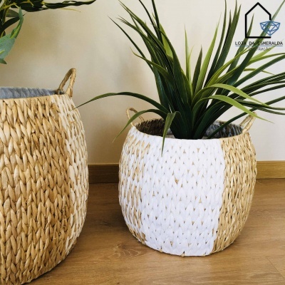 Dois cestos trançados de palha, um com metade pintada de branco, com planta verde dentro, sobre chão de madeira clara