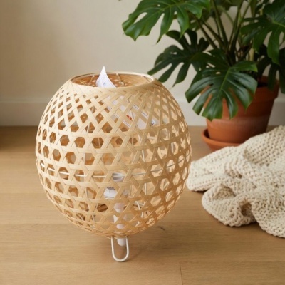Abajur redondo de bambu trançado com base branca ao lado de planta em vaso e manta