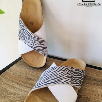 Chinelos com tiras brancas e padrão riscado preto e branco, sobre fundo de madeira com parede branca ao fundo