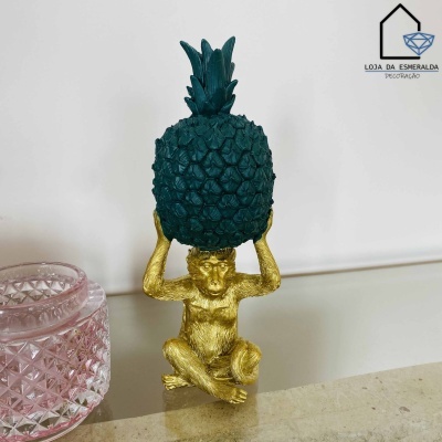 Figura decorativa de macaco dourado com abacaxi verde escuro ao lado de recipiente rosa