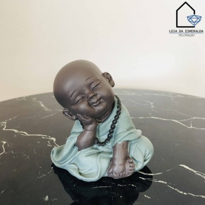 Estátua de monge sorridente sentado com roupa verde clara e colar preto sobre mesa de mármore preto.