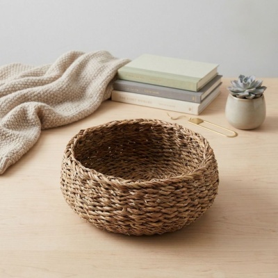 Cesto redondo tecido em fibra natural castanha num ambiente com mesa, cobertor, livros, clipe dourado e vaso com suculenta