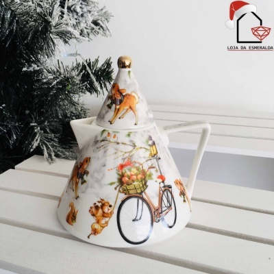 Bule de cerâmica decorado com desenhos de bicicletas e animais, em mesa branca junto a ramo de pinheiro