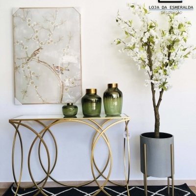 Mesa consola dourada com vasos verdes e quadro floral ao lado de vaso cinzento com flores brancas sobre tapete preto.