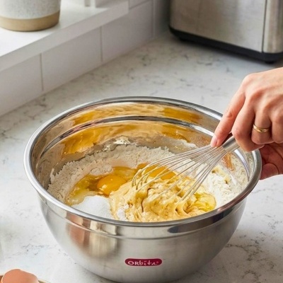 Tigela de aço inoxidável com ingredientes para fazer massa e batedor de arames numa bancada de cozinha.