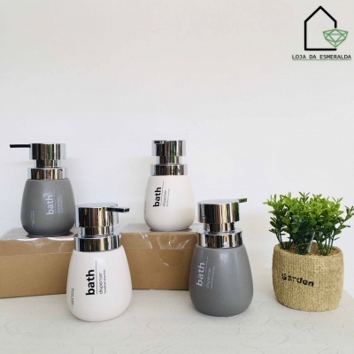 Quatro dispensadores de sabonete em cinza e branco com tampa prateada ao lado de vaso de planta artificial