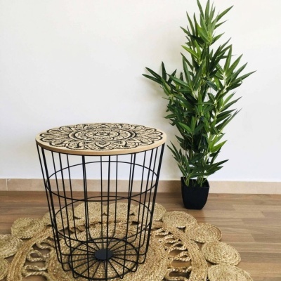 Mesa redonda de madeira com padrão mandala preto e estrutura de metal preto, sobre tapete trançado, ao lado de planta em vaso preto
