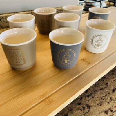 Nove chávenas de café decorativas em cerâmica em diferentes tons, sobre uma tábua de madeira clara.