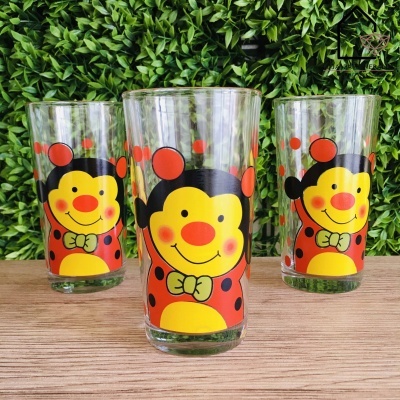 Três copos de vidro com desenhos de joaninhas coloridas sobre madeira e folhagem verde