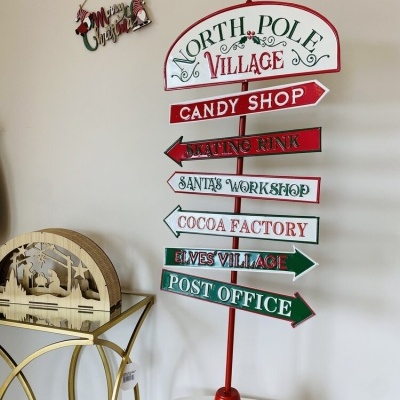 Placa decorativa de Natal com setas indicativas e textos em inglês, suporte vermelho, em mesa branca junto a consola dourada e parede clara.