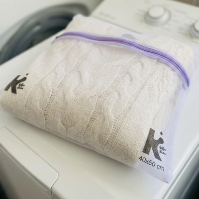 Saco de malha branco com zip violeta e camisola bege dentro, sobre máquina de lavar roupa branca.