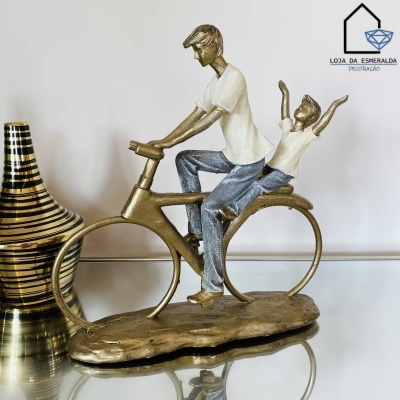 Estatueta de duas figuras humanas numa bicicleta em metal dourado sobre superfície de vidro ao lado de vaso decorativo