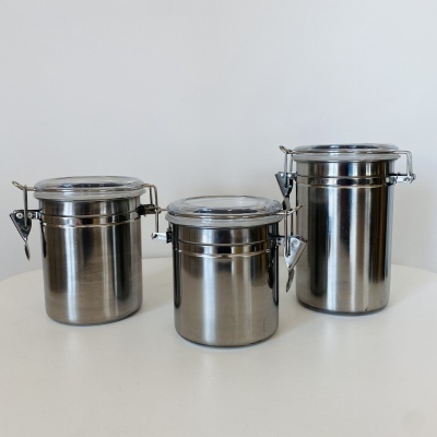 Três recipientes cilíndricos de aço inox com tampas transparentes e fechos metálicos