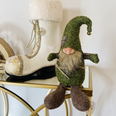 Bota decorativa branca com pelúcia e gnomo decorativo verde sentados numa mesa dourada com tampo de vidro