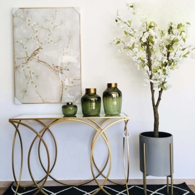 Mesa de entrada dourada com vidro e decoração com jarras verdes, quadro de pássaros e planta artificial branca