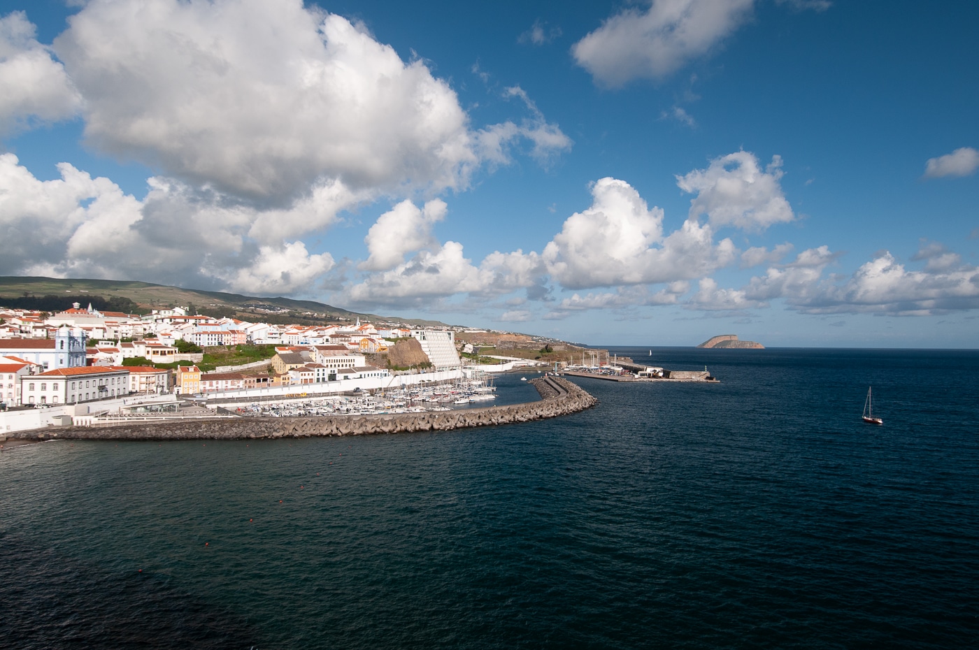 Vista costeira de cidade com porto, edifícios brancos, céu azul e barco à vela