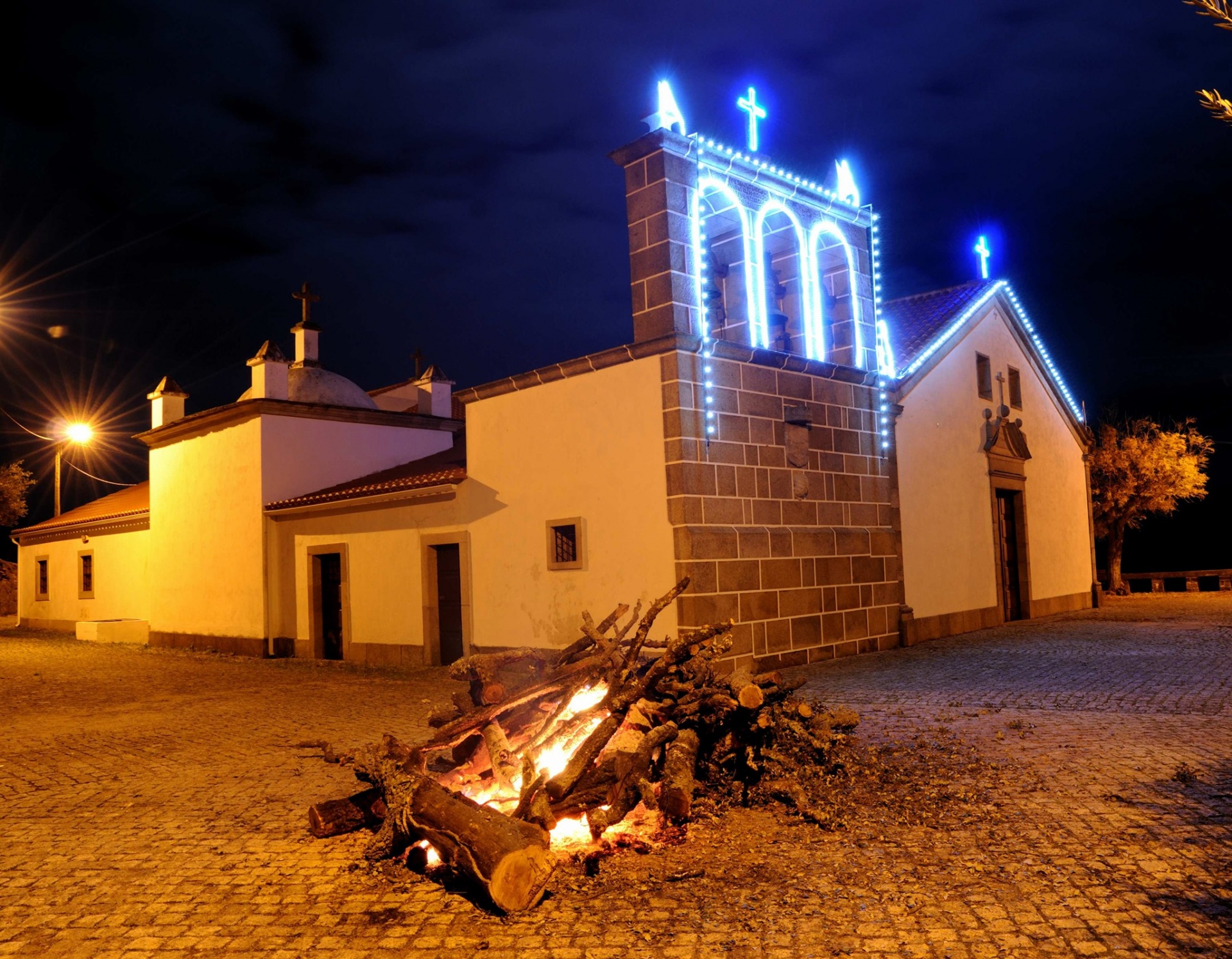 Igreja branca com torre iluminada a azul e fogueira em primeiro plano à noite