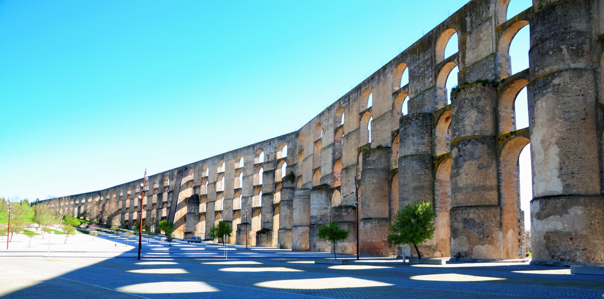 Aqueduto antigo com várias arcadas em pedra amarelada junto a estrada com sombra