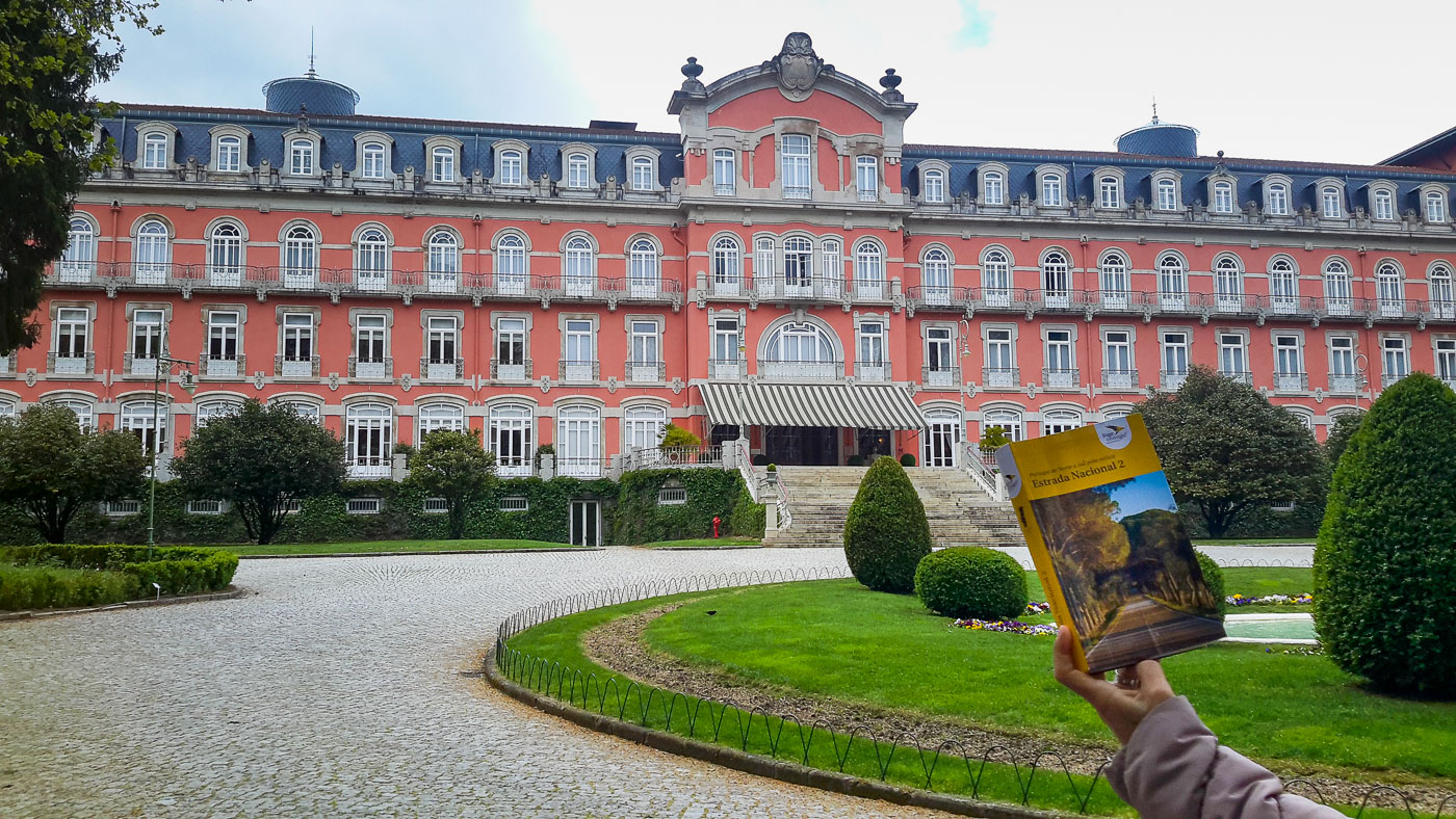 Edifício antigo rosa com janelas brancas, jardim e caminho em pedras, mão segura guia turístico amarelo