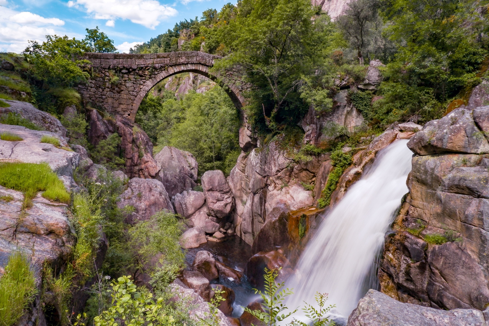 Ponte de pedra em arco sobre desfiladeiro com cascata e vegetação