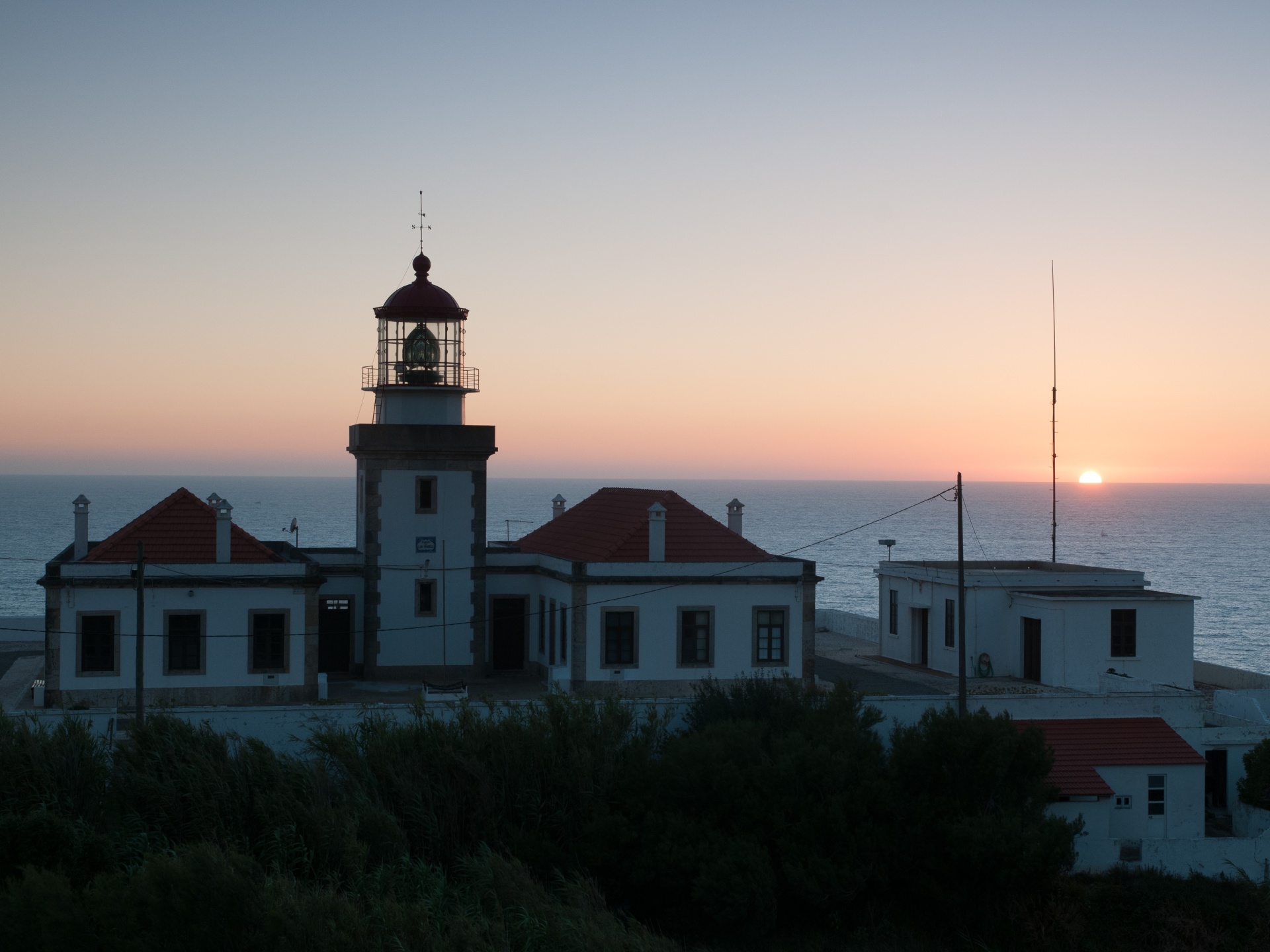 Farol branco com molduras pretas e telhados vermelhos junto ao mar ao pôr do sol