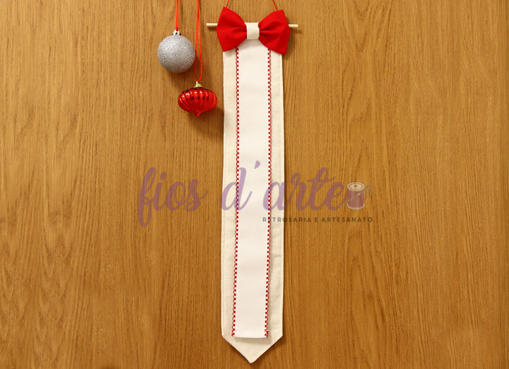 Gravata bege com bordado vermelho e laço vermelho pendurada numa parede de madeira com bolas de Natal ao lado