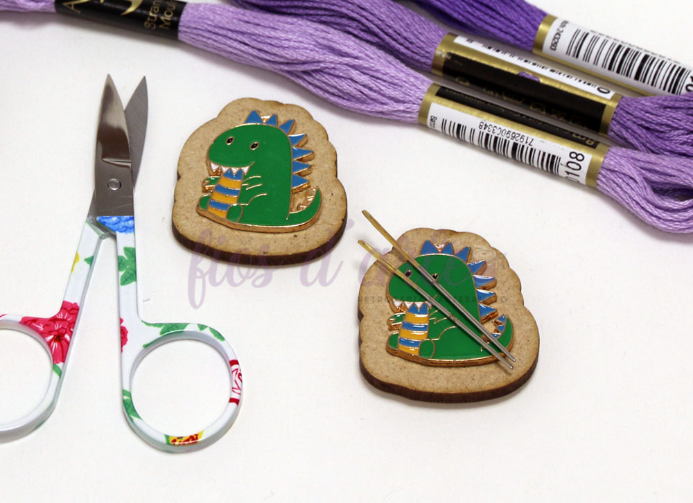 Suporte magnético para agulhas - Dinossauro Alfinetes de dinossauro em base castanha com agulhas, tesoura floral e novelos de linha roxa.