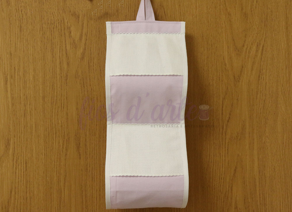 Suporte Liso Rolo Papel Higiénico para bordar ponto cruz (Fios d'arte) Suporte em tecido branco e lilás para rolo de papel pendurado numa parede de madeira.