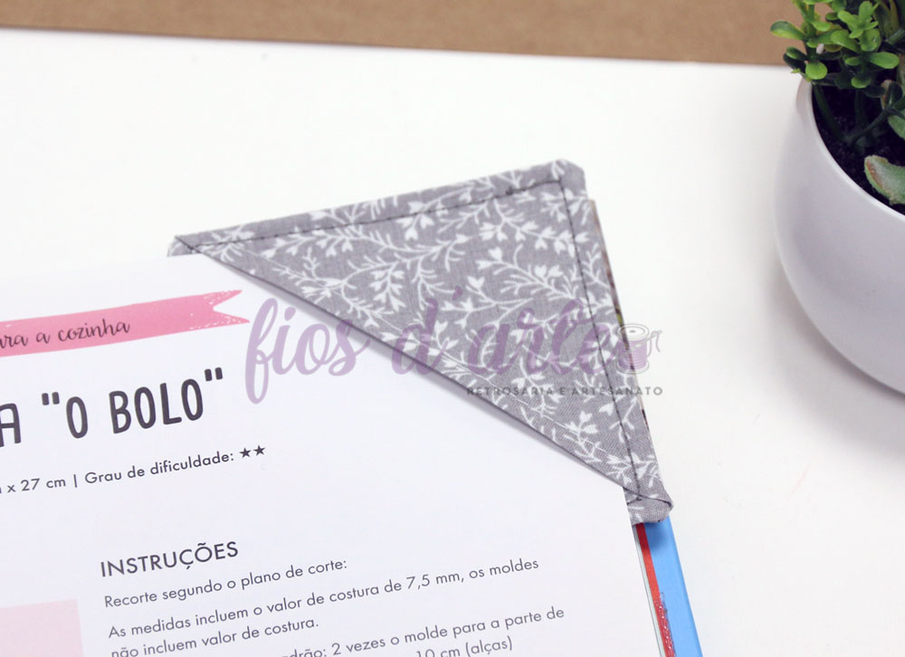 Marcador de livro triangular cinza com padrão branco sobre página de livro e vaso de planta