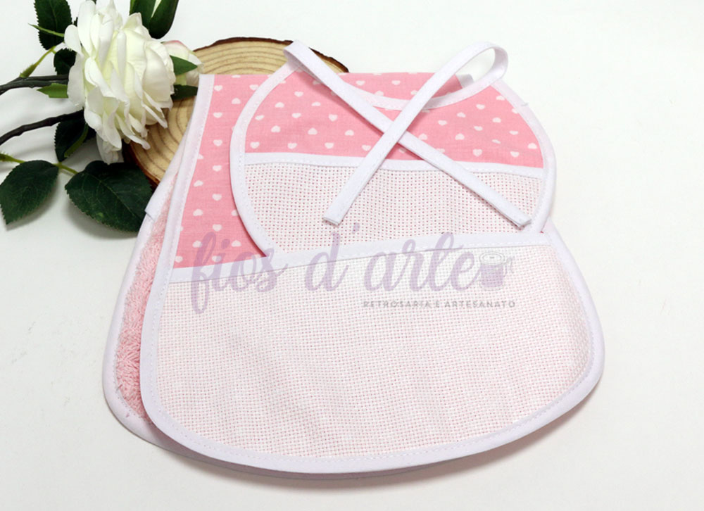 Conjunto de babete rosa com padrão de corações brancos e toalha rosa ao lado de flor branca e madeira