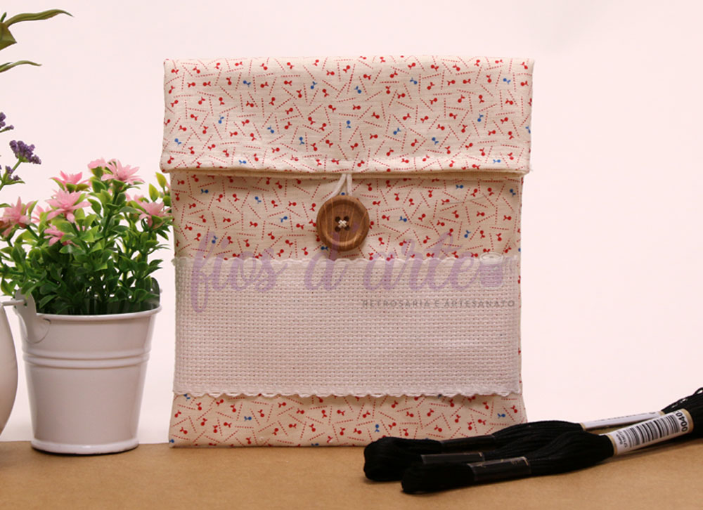 Estojo em tecido bege com padrão floral, botão de madeira e detalhe branco em mesa com planta e linhas