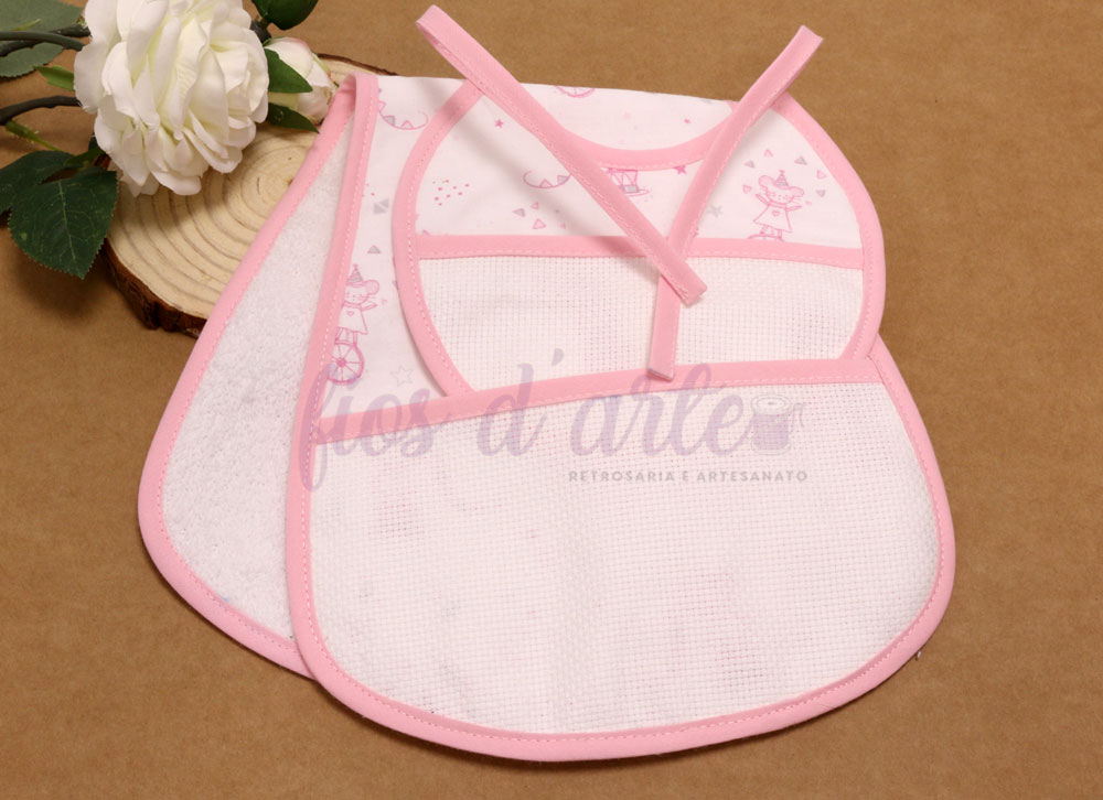 Conjunto de três babetes de bebé brancos com bordas e desenhos cor-de-rosa, sobre fundo castanho com flor branca e base de madeira.