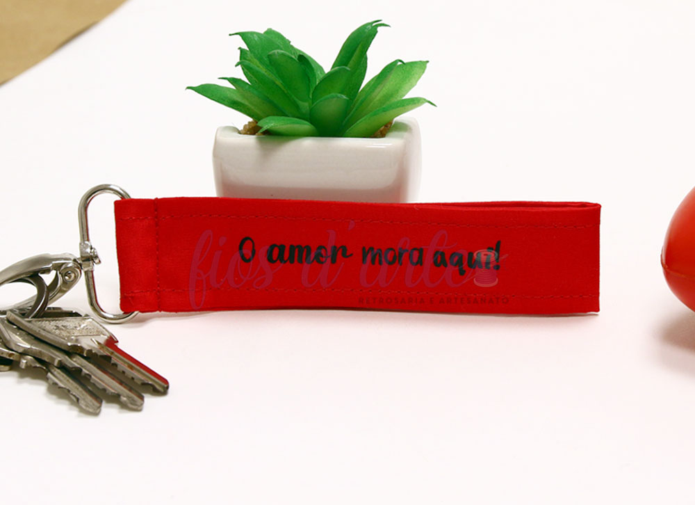 Chaveiro vermelho com texto 'O amor mora aqui' e planta artificial ao fundo