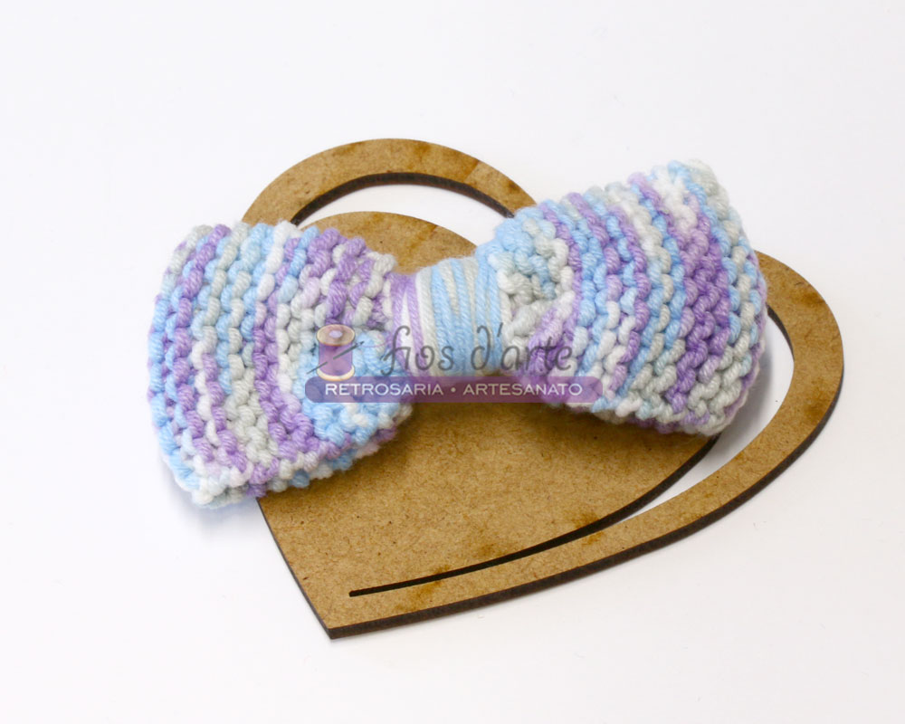 Laço de cabelo tricotado azul, branco e lilás sobre coração de madeira.