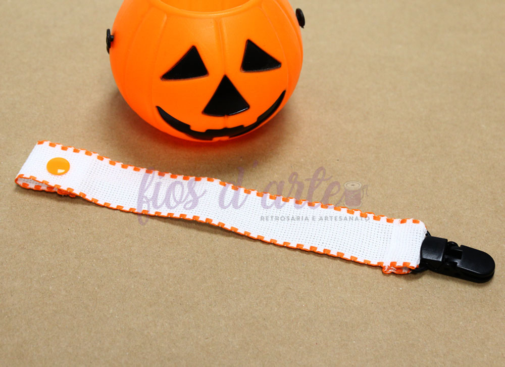 Prende-chupeta com fita em quadrilé para bordar ponto cruz #15 (Fios d'arte) 🎃 Pulseira para bebé branca com bordas laranja e fecho preto em superfície castanha ao lado de balde de abóbora laranja de Halloween