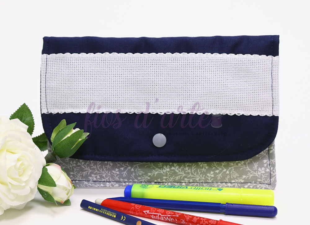 Estojo azul escuro com aba branca e botão, canetas coloridas e flores brancas