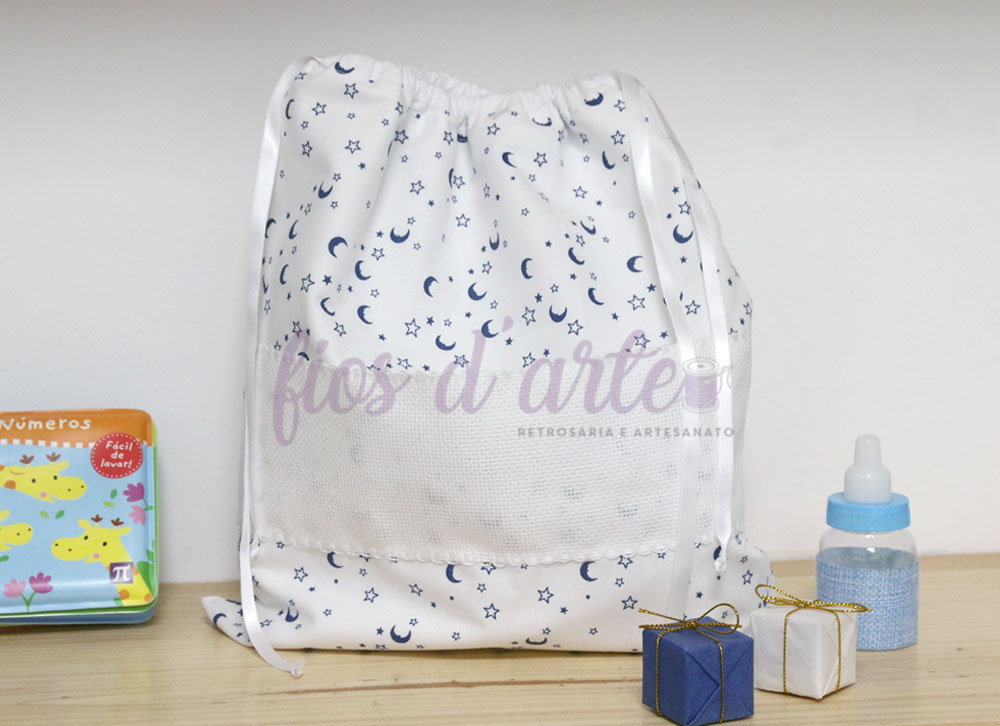 Bolsa/ Saco forrado 1ª roupinha para bordar ponto cruz (Fios d'arte) Saco de tecido branco com estampado azul de luas e estrelas, com cordões de cetim, sobre mesa de madeira com acessórios infantis