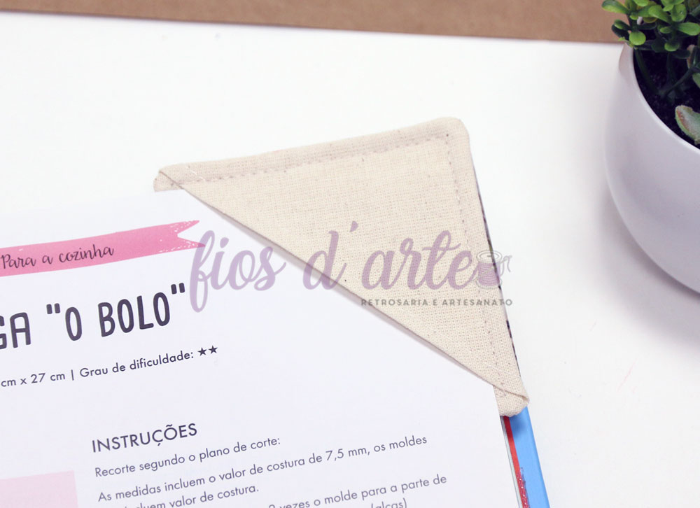 Pano de cozinha triangular bege sobre folha de instruções em português junto a planta em vaso branco