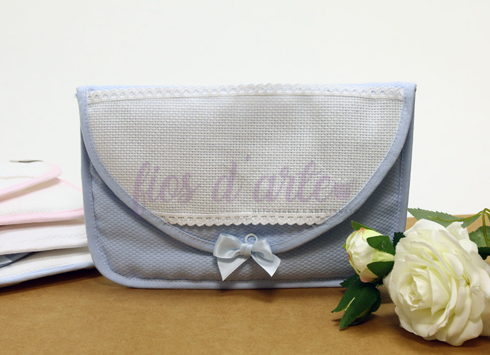 Carteira cinzenta e branca com bordado, renda e laço, sobre mesa castanha com flores brancas