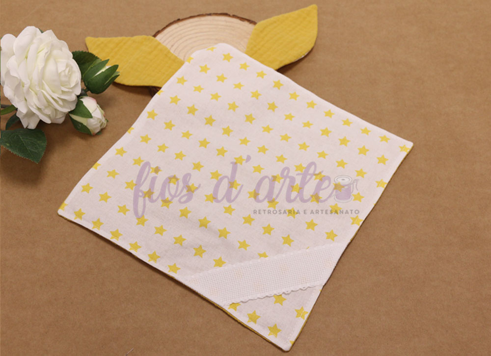 Toalha branca com estrelas amarelas e orelhas amarelas, junto a uma flor branca sobre fundo castanho.