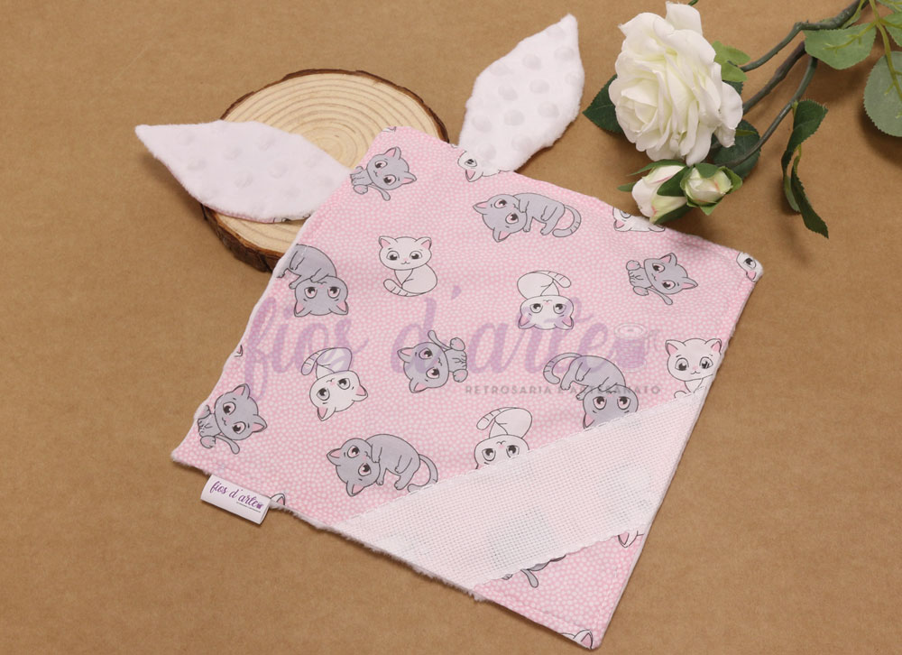 Manta bebé rosa com padrão de gatos e orelhas brancas felpudas junto a flores brancas e base de madeira