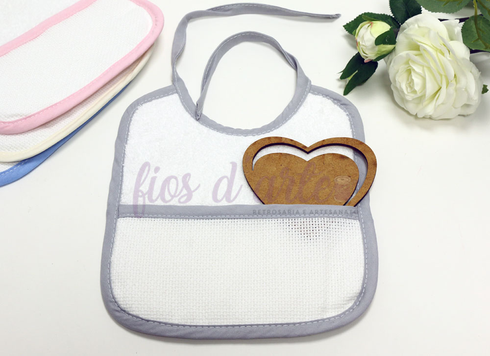 Babete branco com bordas cinzentas e bolso frontal com desenho de pães castanhos, vários babetes coloridos ao fundo e flores brancas ao lado.