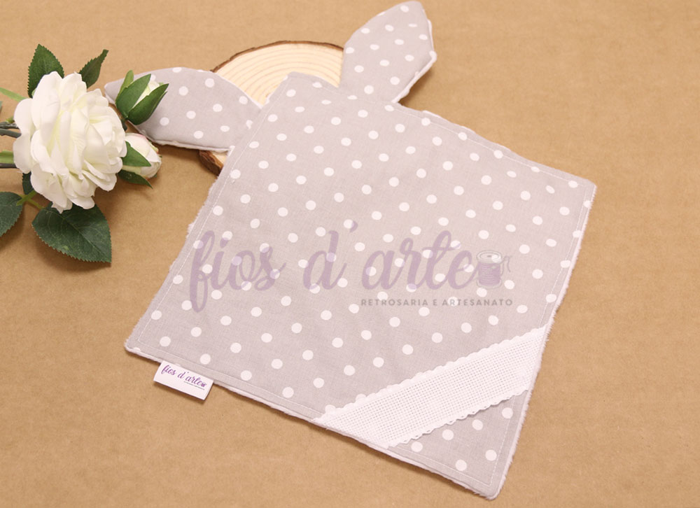 Toalha cinza com bolinhas brancas em forma de coelho com orelhas, sobre fundo castanho claro com flor branca ao lado