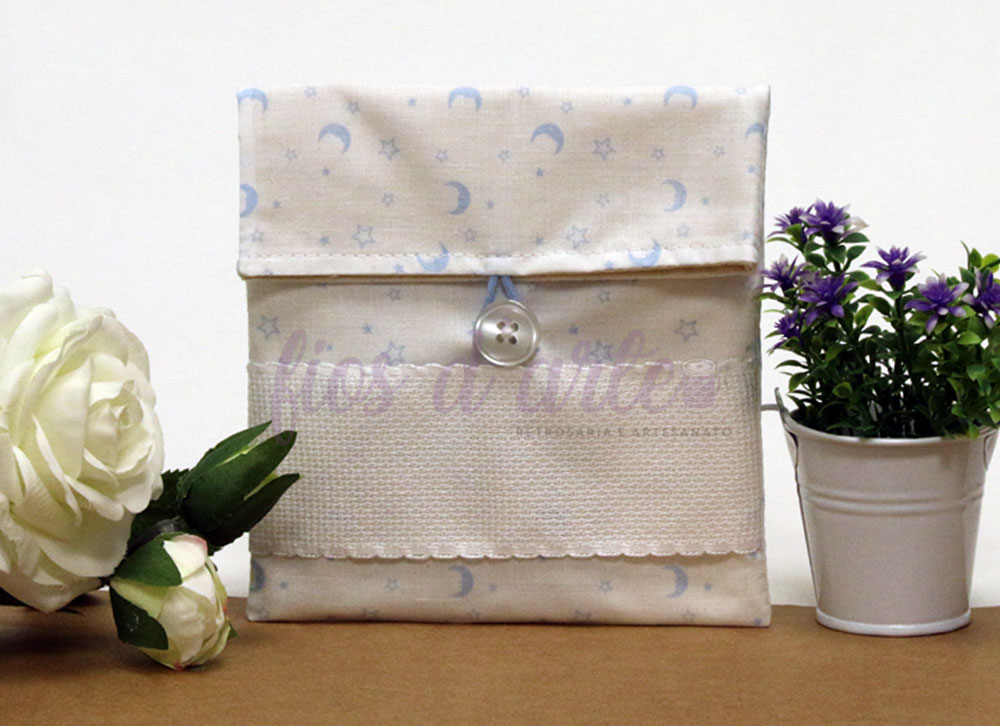 Bolsa Multiusos estampada com quadrilé para bordar ponto cruz (14x15cms) - Estrelas e Luas Bolsa de tecido com padrão de luas e estrelas azuis em fundo bege, detalhe em renda branca, entre flores artificiais branca e púrpura.