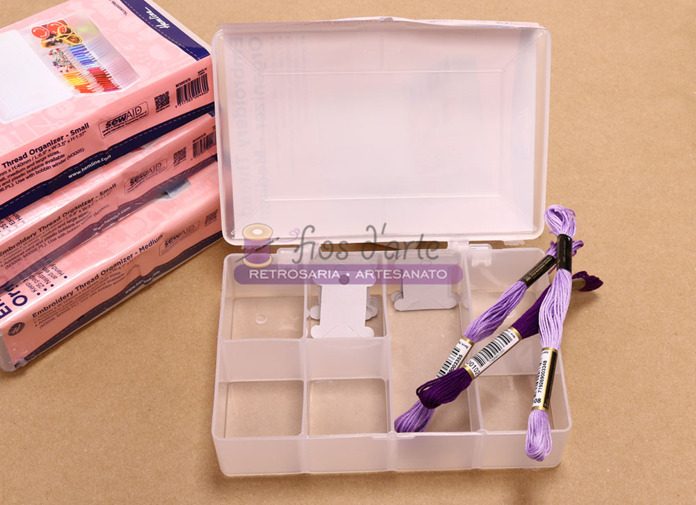 Caixa organizadora para fios com bobines de cartão (Hemline) - média Caixa plástica transparente com linhas de bordado roxas em divisórias à superfície castanha e embalagens cor-de-rosa ao lado